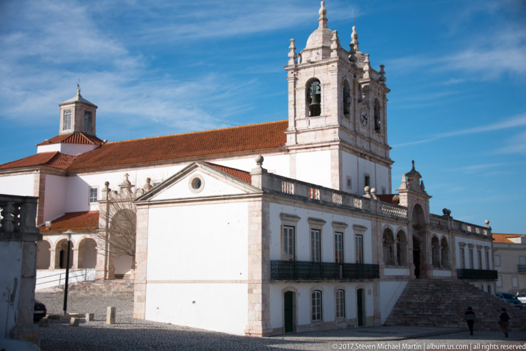 White church complex with terracotta roofs, stone stairway, and tall bell tower under blue sky.
