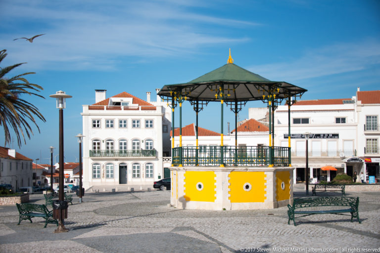 Yellow-and-white octagonal bandstand with metal railings in a paved Nazaré square, surrounded by benches and white buildings.