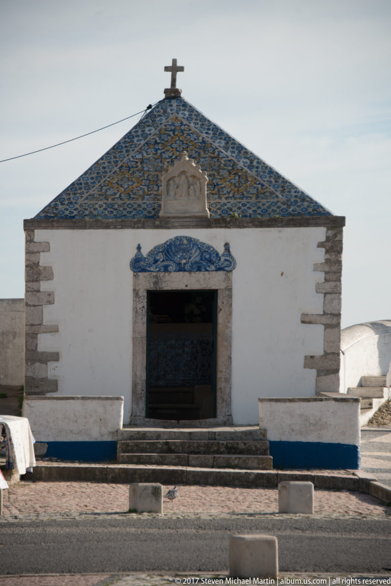 Small white chapel with stone doorway, blue-and-white tiled pediment, corner stones, steps, and a cross at the roof peak.