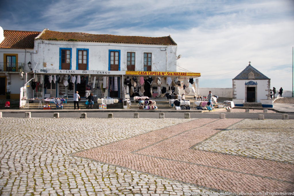 Souvenir shops and outdoor merchandise beside a small chapel across a broad patterned cobblestone square in Nazaré.