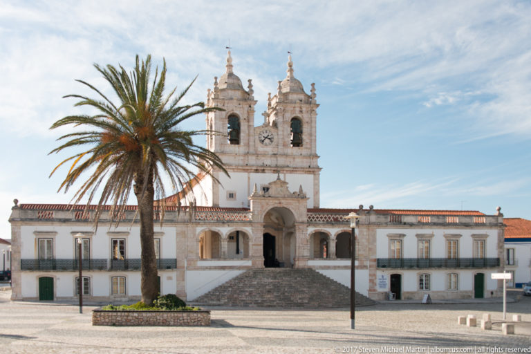 Front view of the Sanctuary of Our Lady of Nazaré with twin bell towers, central staircase, arched entrance, and a tall palm.
