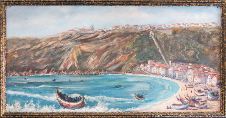 Framed painting of Nazaré bay with beach, cliff-top settlement, waterfront buildings, surf, and traditional boats in the foreground.