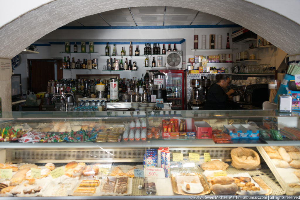 Glass pastry counter filled with baked goods in a small Nazaré café, with bottles, coffee equipment, and a worker behind the bar.