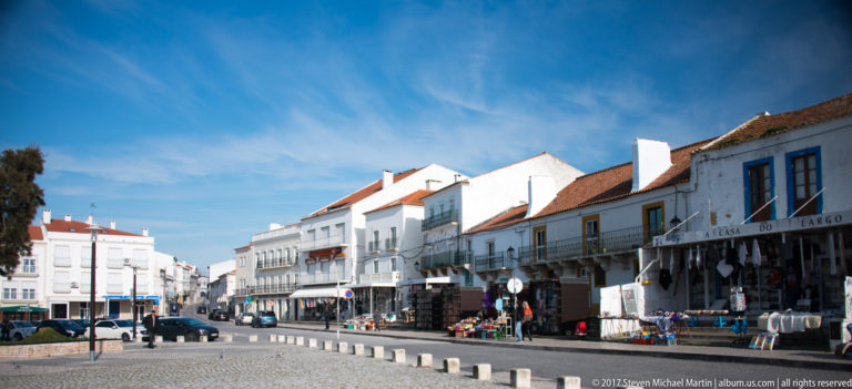 Curving Nazaré street lined with white buildings, balconies, red tile roofs, souvenir displays, bollards, and parked vehicles.