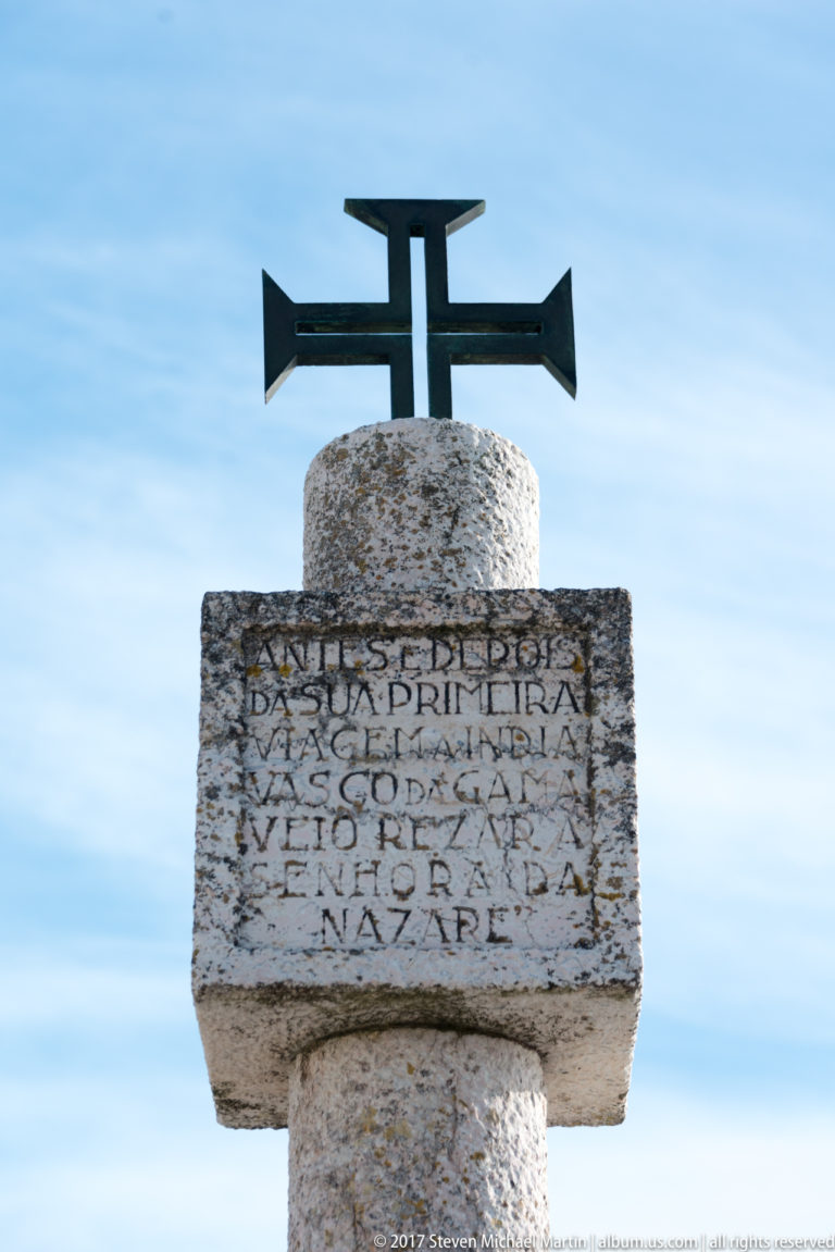 Weathered stone monument topped by a dark metal cross, with a carved Portuguese inscription ending in the word Nazaré.