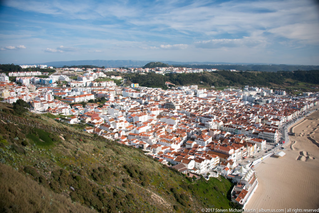 Elevated view from Nazaré's cliff across dense white-roofed town blocks toward the sandy Atlantic beachfront.