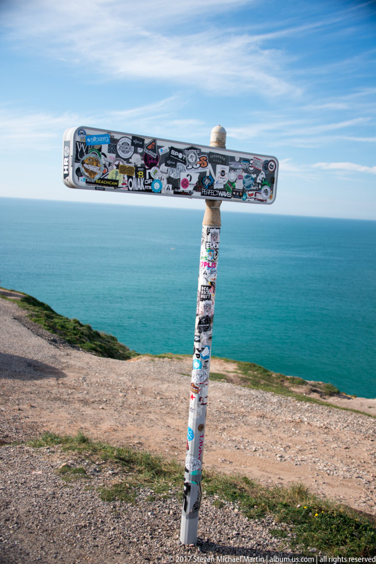 Sticker-covered directional signpost standing on a headland above the blue Atlantic Ocean.