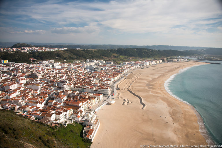 Wide cliff-top panorama of Nazaré's curved sandy beach, blue Atlantic water, and dense lower town along the shoreline.