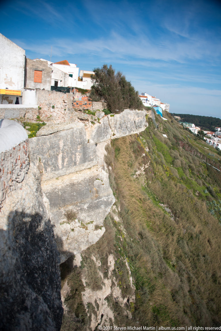 Weathered masonry and exposed pale rock on Nazaré's clifftop, with scrubby slope and white buildings beyond.