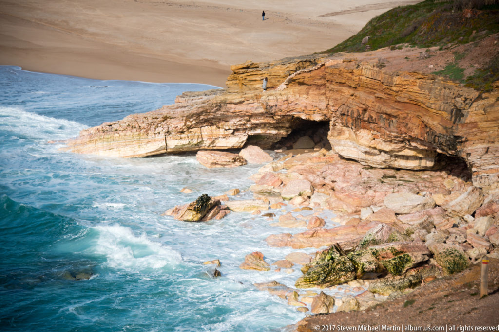 White surf washing around layered orange rock ledges and a dark sea cave at the cliff base.