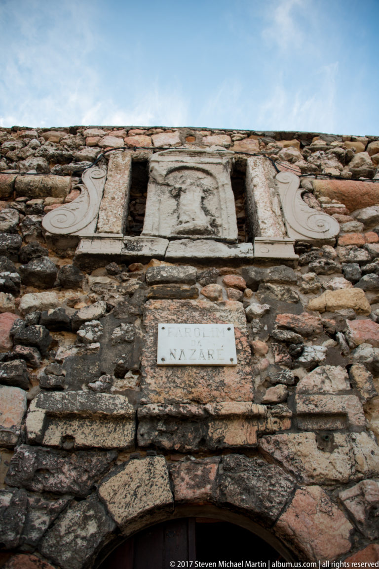 Arched doorway in a rough stone wall beneath carved ornament and a plaque reading Nazaré.