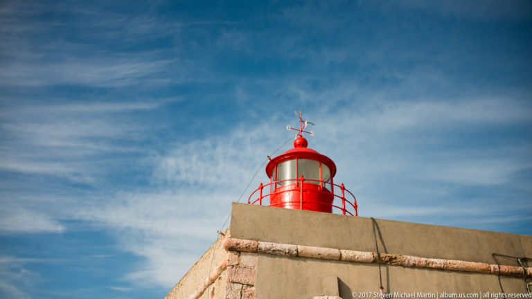Bright red lighthouse lantern and gallery rising above pale masonry under a blue sky.