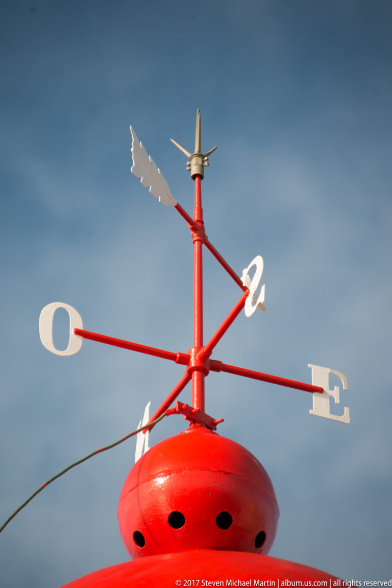 Red lighthouse cap topped by a weather vane with white cardinal-direction letters against blue sky.