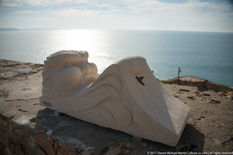 Pale carved stone fragment with curling forms resting on a masonry ledge above the ocean.