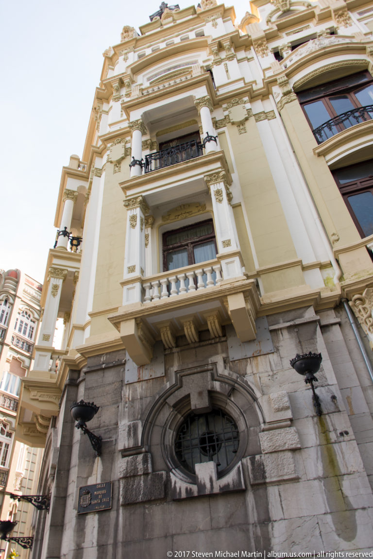 Upward view of an ornate cream-colored façade with stone base, oval window, balconies, pilasters, and sculpted architectural details.