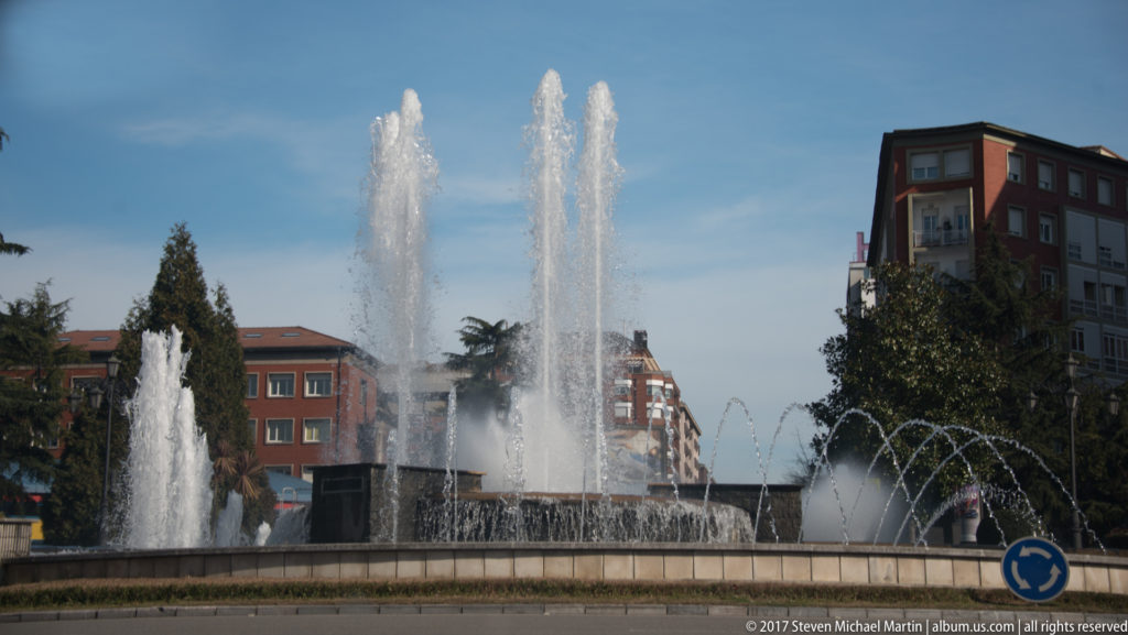 Circular urban fountain with tall vertical jets and smaller arcing sprays, surrounded by buildings, trees, and a roundabout sign.