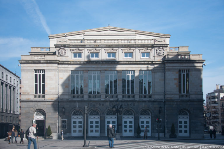 Symmetrical stone façade of Teatro Campoamor with pediment, upper windows, entrance doors, and pedestrians crossing the plaza.