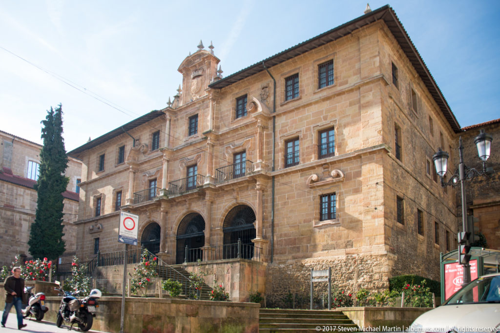 Large honey-colored stone building with three entrance arches, rows of windows, carved shields, and a central crest above the façade.