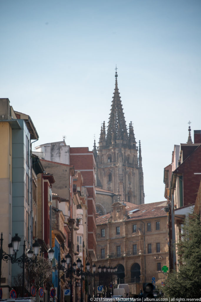 Gothic spire of Oviedo Cathedral rising above a straight city street lined with buildings, roofs, and street lamps.