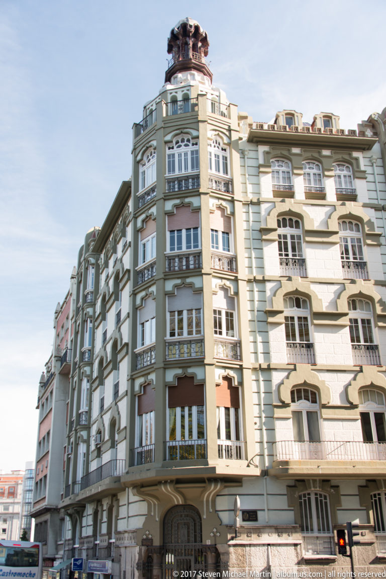 Rounded multi-story corner building with bay windows, balconies, pale green and cream façades, and a small rooftop turret.