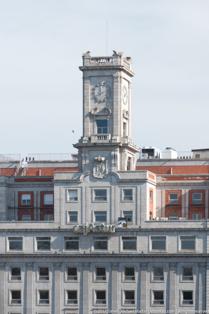 Frontal view of a square stone civic clock tower above a symmetrical façade with a sculpted heraldic emblem.