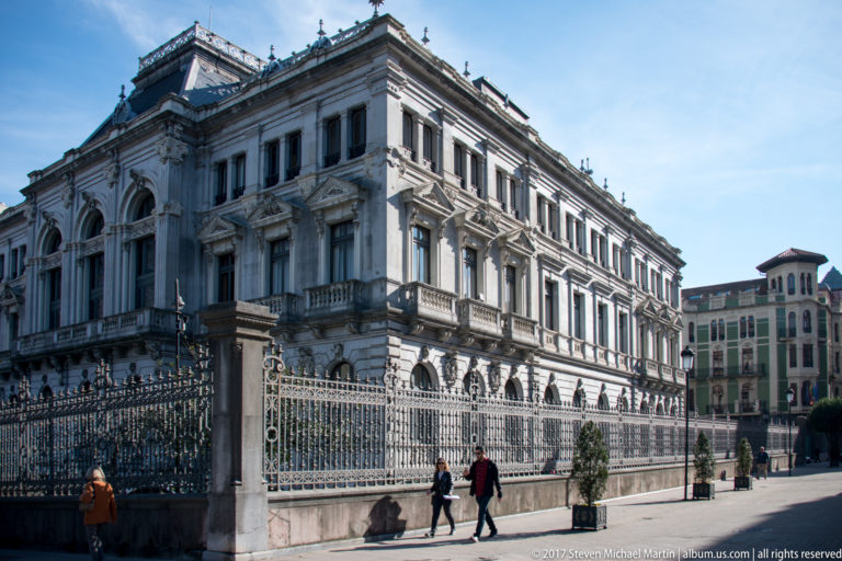 Long stone façade of the historic University of Oviedo seen behind ornate wrought-iron fencing, with pedestrians on the sidewalk.