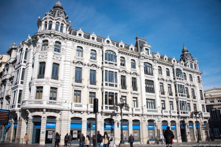 Large ornate white corner building with curved bays, balustrades, dormers, dark roof caps, storefronts, and pedestrians below.