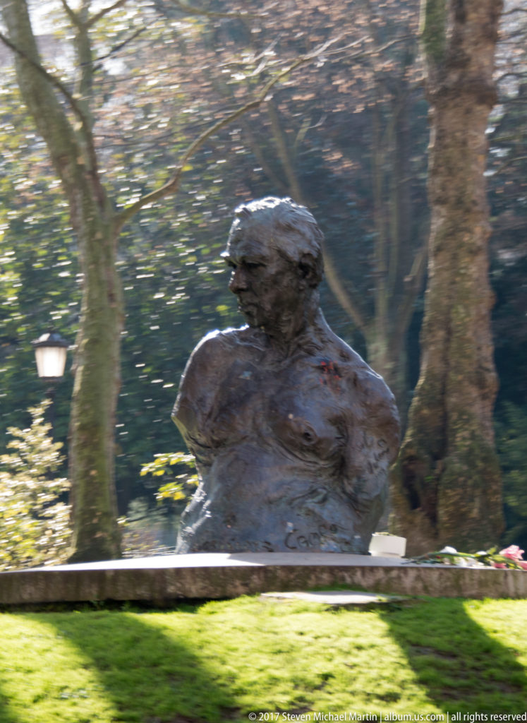 Close view of a textured bronze human figure among trees and lawn in a shaded city park.