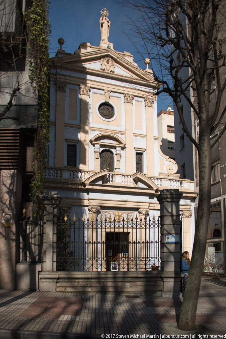 Symmetrical cream chapel façade with arched doorway, oval window, pediment, and small rooftop finial behind an iron gate.