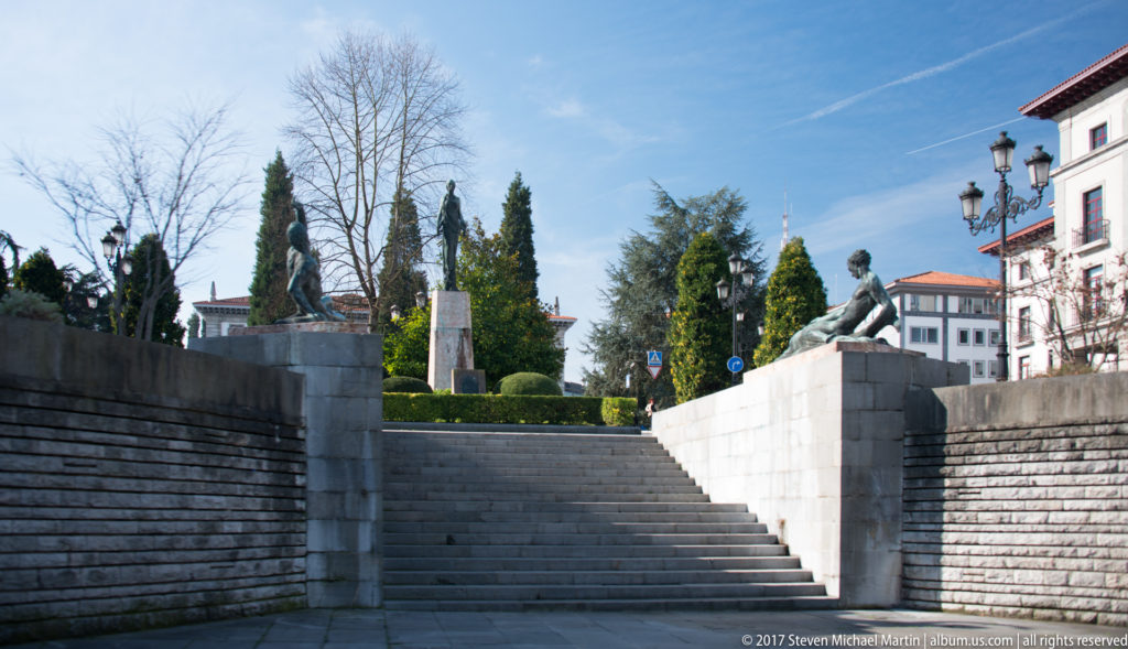 Wide stone steps rise between retaining walls toward trees, street lamps, and a reclining bronze figure on the upper parapet.