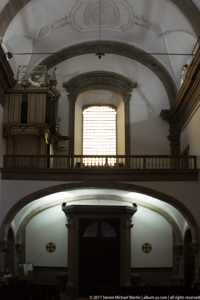 Church entrance interior with broad arch, upper gallery, large window, and dark wooden organ case at left.