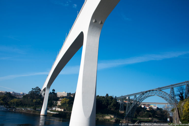 White concrete Ponte do Infante curves overhead, with the iron Maria Pia Bridge spanning the Douro in the distance.