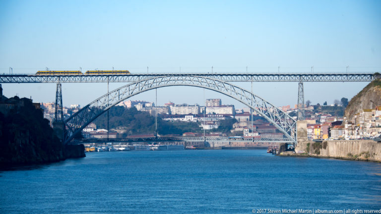 Yellow train crosses the high deck of an iron arch bridge spanning the Douro between steep banks.