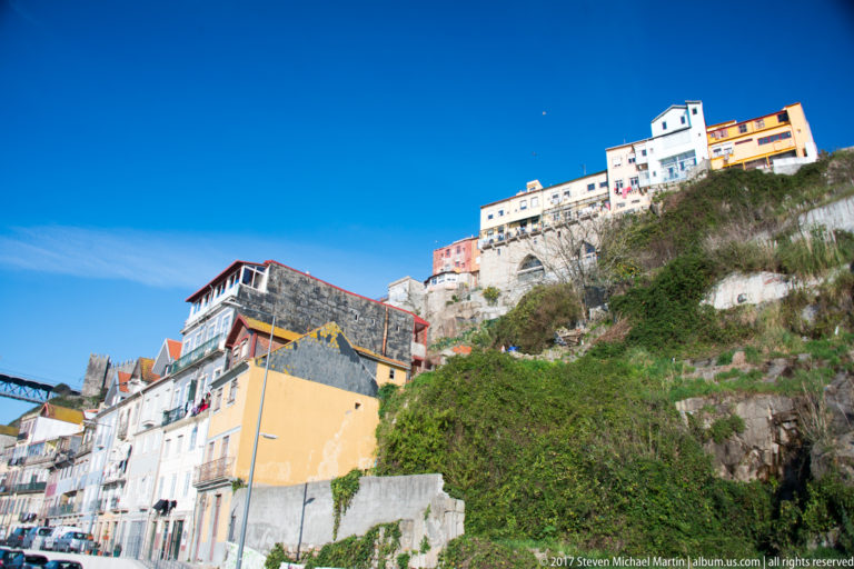 White and pastel houses step up a rocky green hillside beneath a clear blue sky in Porto.