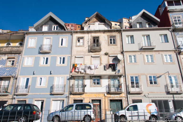 Narrow pastel and stone residential façades with small balconies, laundry and parked cars along a Porto street.