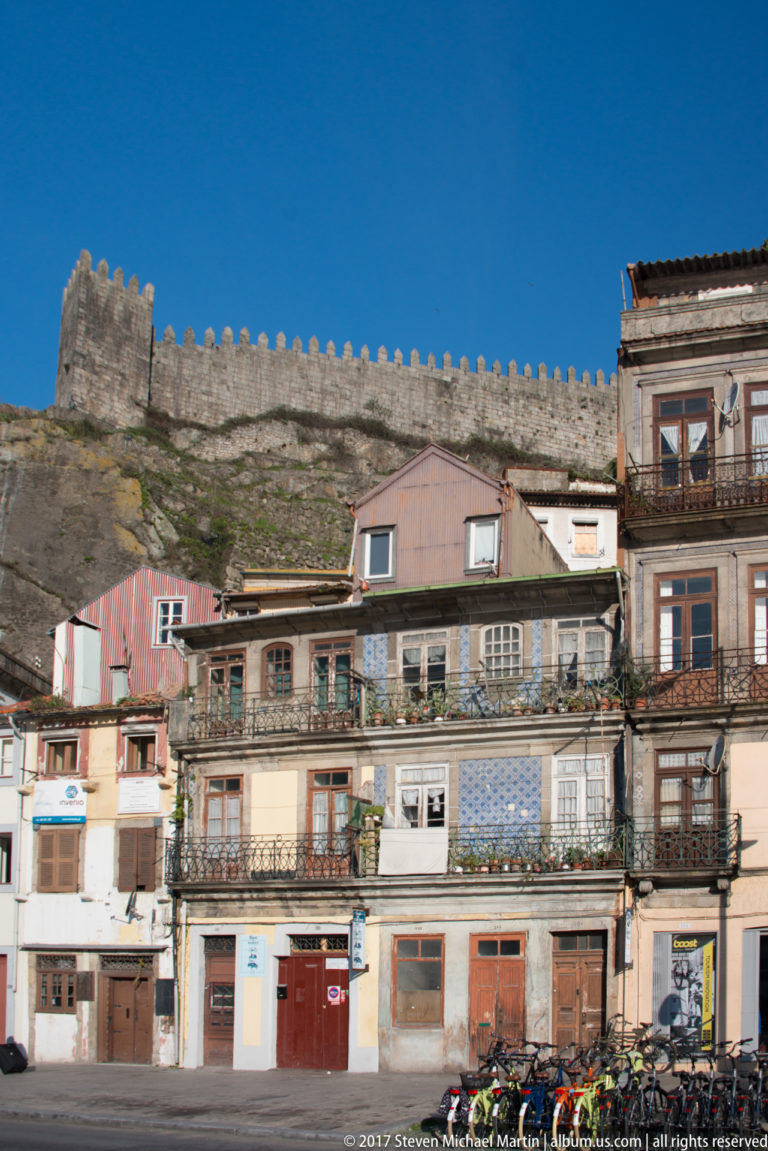 Crenellated stone fortification wall runs along a hillside above narrow residential buildings with balconies and tiled façades.