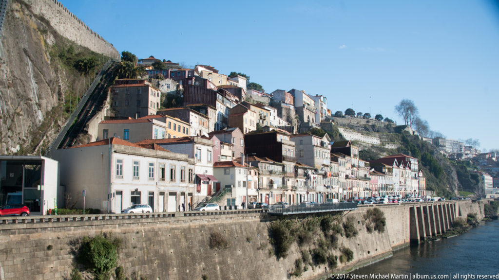 Road and houses follow a stone embankment beneath a steep, densely built Porto hillside beside the Douro.