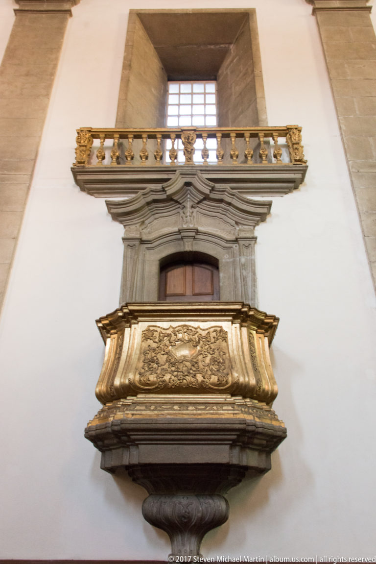 Projecting carved stone tribune with balustrade, doorway, and high window against a white church wall in Porto.