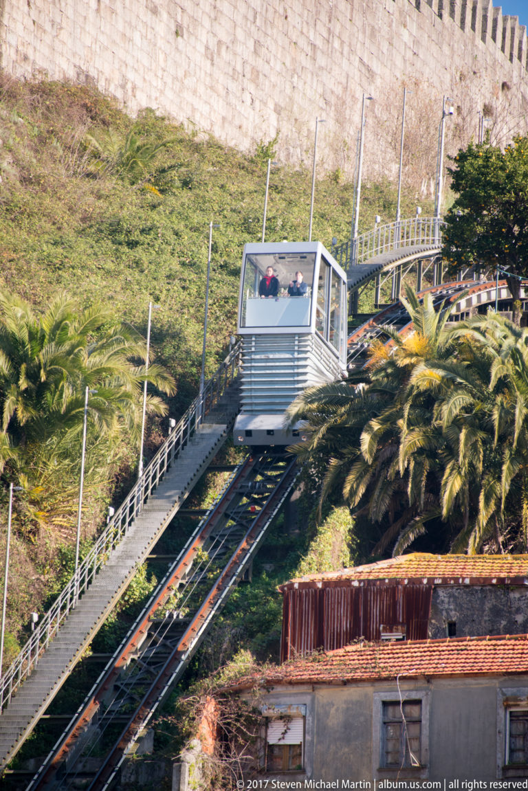 Silver Funicular dos Guindais car climbs steep rails through a green slope below a high stone wall in Porto.