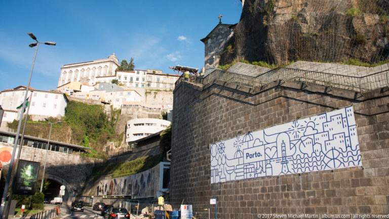 Blue-and-white mural reading “Porto.” decorates a dark stone retaining wall beneath steep upper-city buildings.