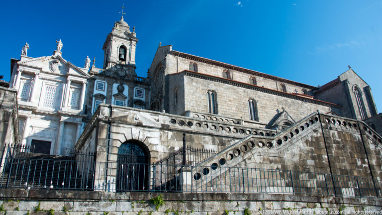 Large stone church with exterior stair, arched doorway and adjoining ornate white façade and bell tower in Porto.