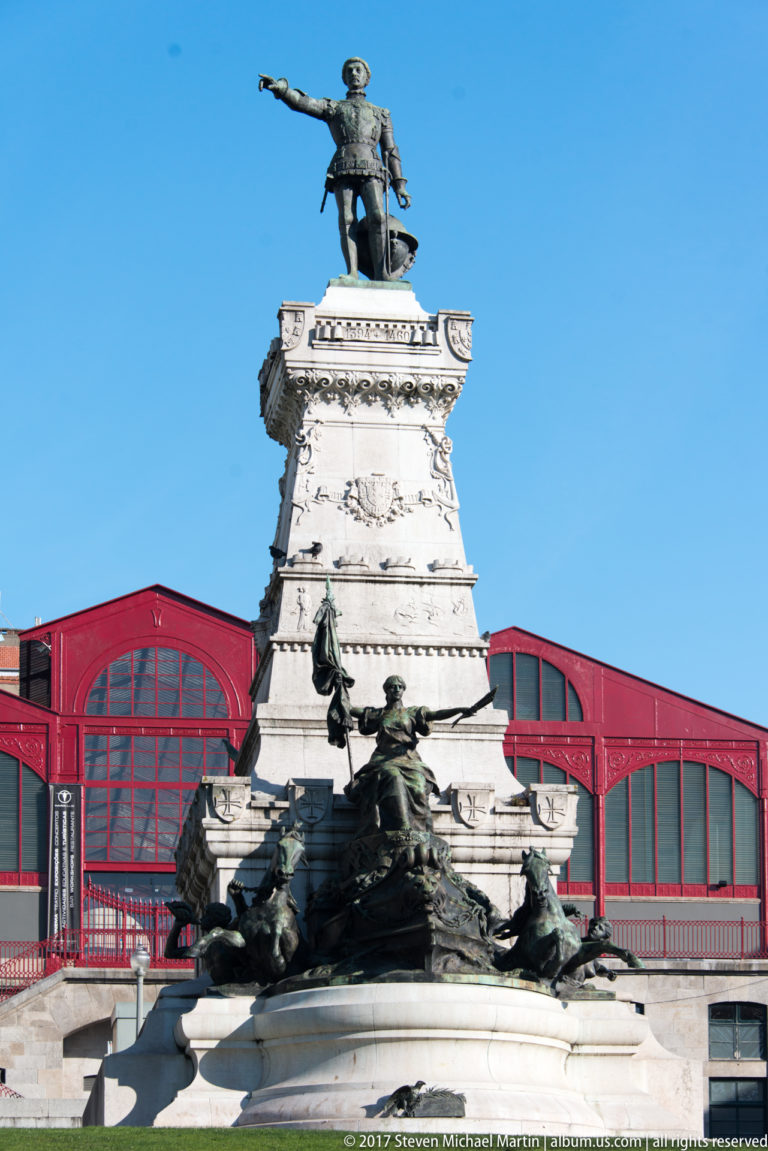 Tall Prince Henry the Navigator monument with bronze figures at its base, backed by the red Mercado Ferreira Borges.