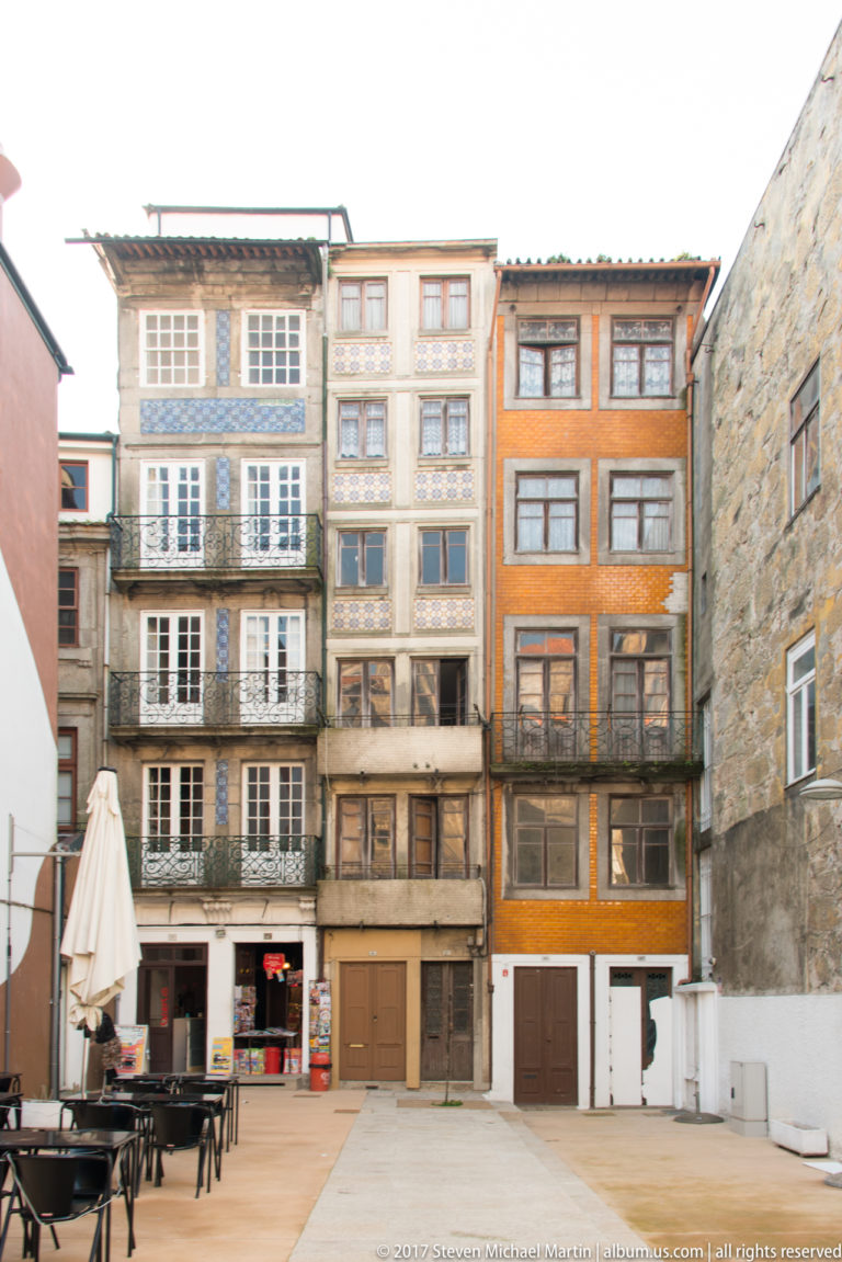 Tall narrow historic façades with stacked windows surround a paved terrace with café tables in Porto.