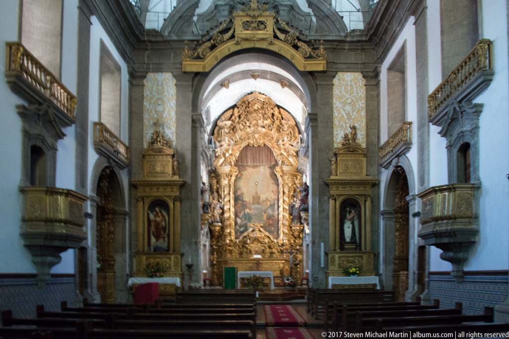 Church nave with wooden pews leading to an elaborate gilded Baroque main altarpiece framed by pale stone arches.