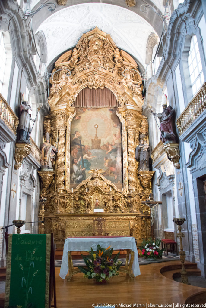 Elaborate gilded Baroque retable surrounds a central religious painting above a white altar, with sculpted figures at both sides.