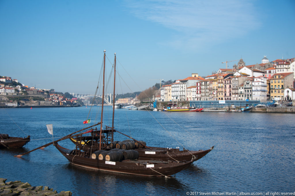 Wooden rabelo boat with wine barrels and a long steering oar floating before Porto’s historic riverfront.