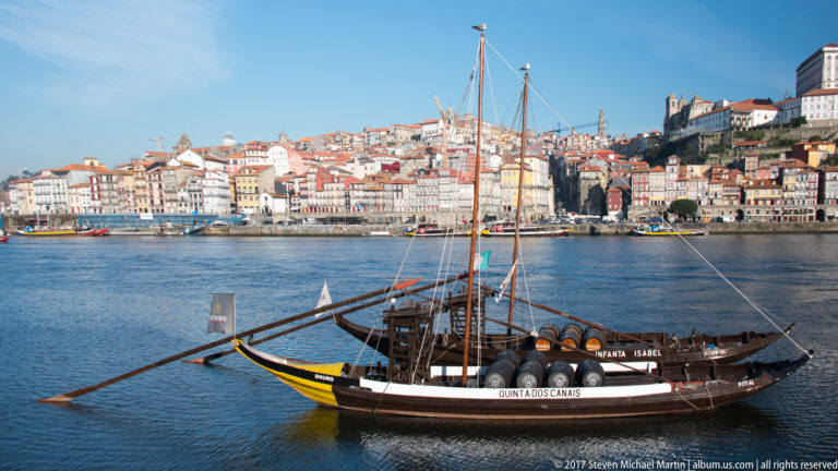 Broadside view of a wooden rabelo boat carrying barrels on the Douro before Porto’s steep historic hillside.