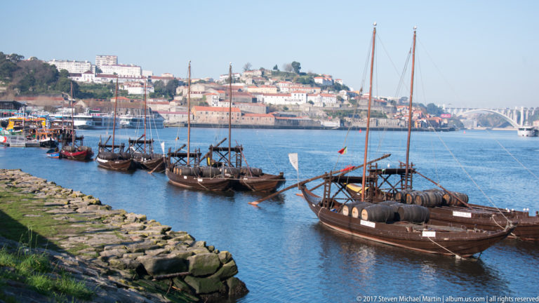 Several wooden rabelo boats moored along a stone bank on the Douro, with Porto’s river corridor beyond.