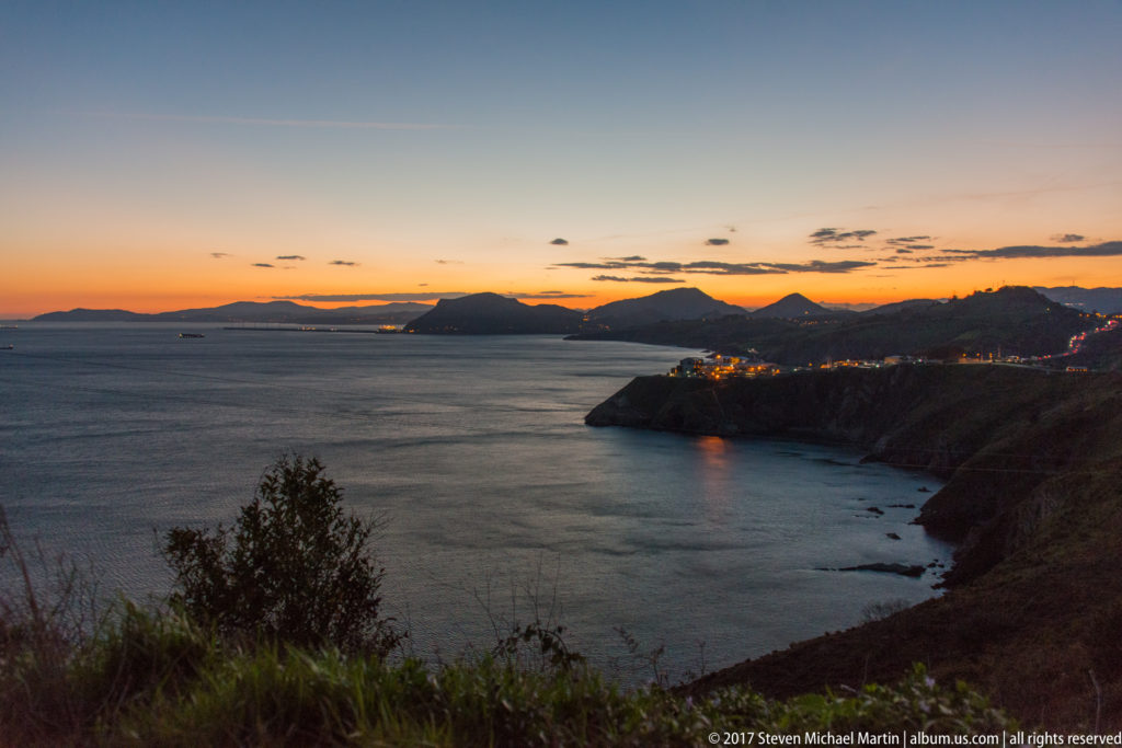 Cantabrian coastline at sunset with dark headlands, calm sea, distant mountains, and scattered shoreline lights.