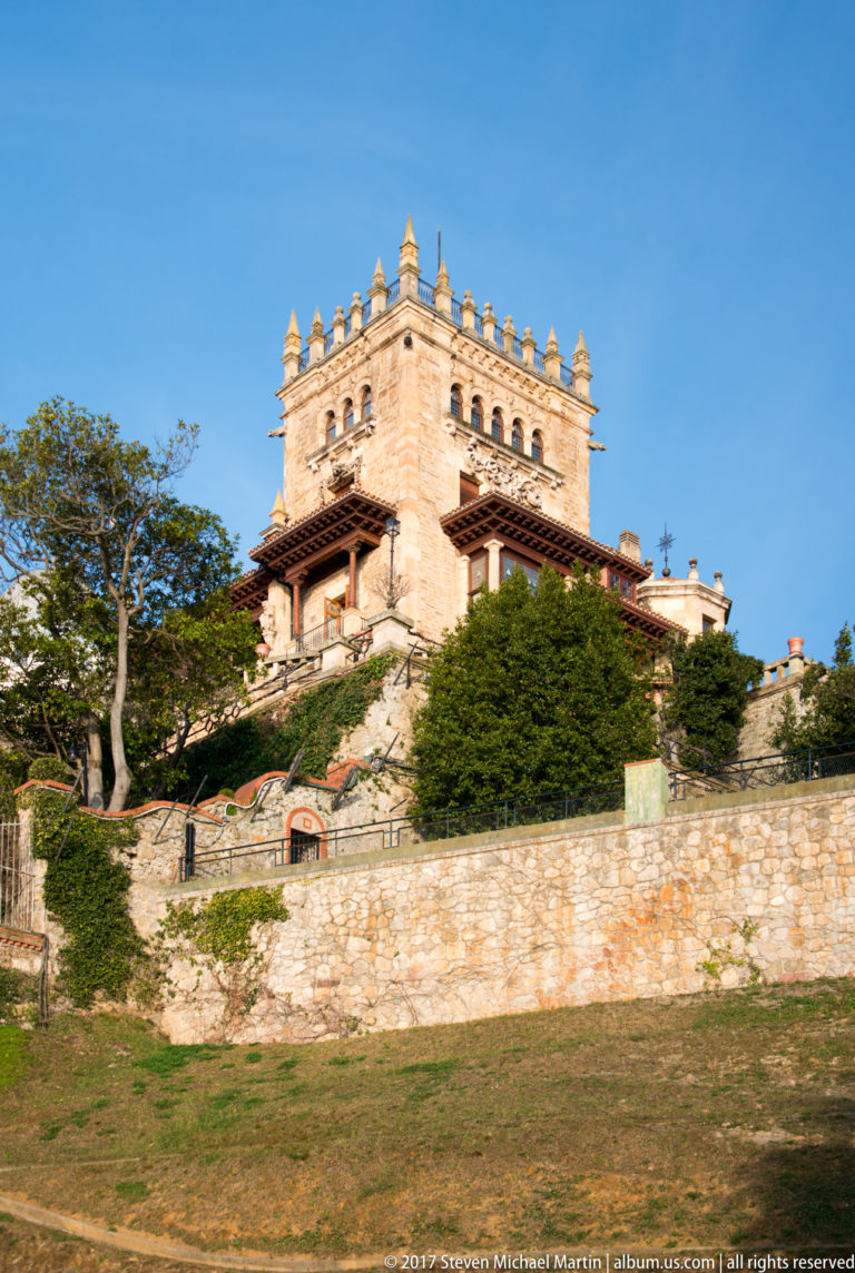 Stone Palacio de la Magdalena on a landscaped rise, with crenellated roofline, timbered eaves, trees, and terrace walls.